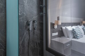 Ein modernes Badezimmer mit Duschkabine aus Glas, einer Marmorwand und Blick in ein Schlafzimmer durch das Glas.