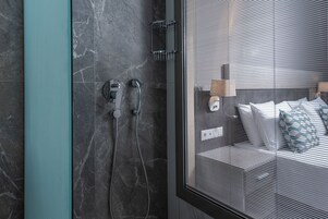 Un baño moderno con cabina de ducha de vidrio, pared de mármol y vista a un dormitorio a través del vidrio.