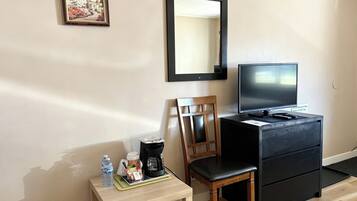 Habitación de hotel con una silla de madera, una mesita con una botella de agua, una cafetera y un televisor sobre un pedestal negro.