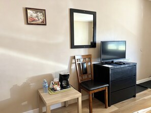 Quarto de hotel com uma cadeira de madeira, uma mesa pequena com uma garrafa de água, uma cafeteira e uma televisão sobre um suporte preto.