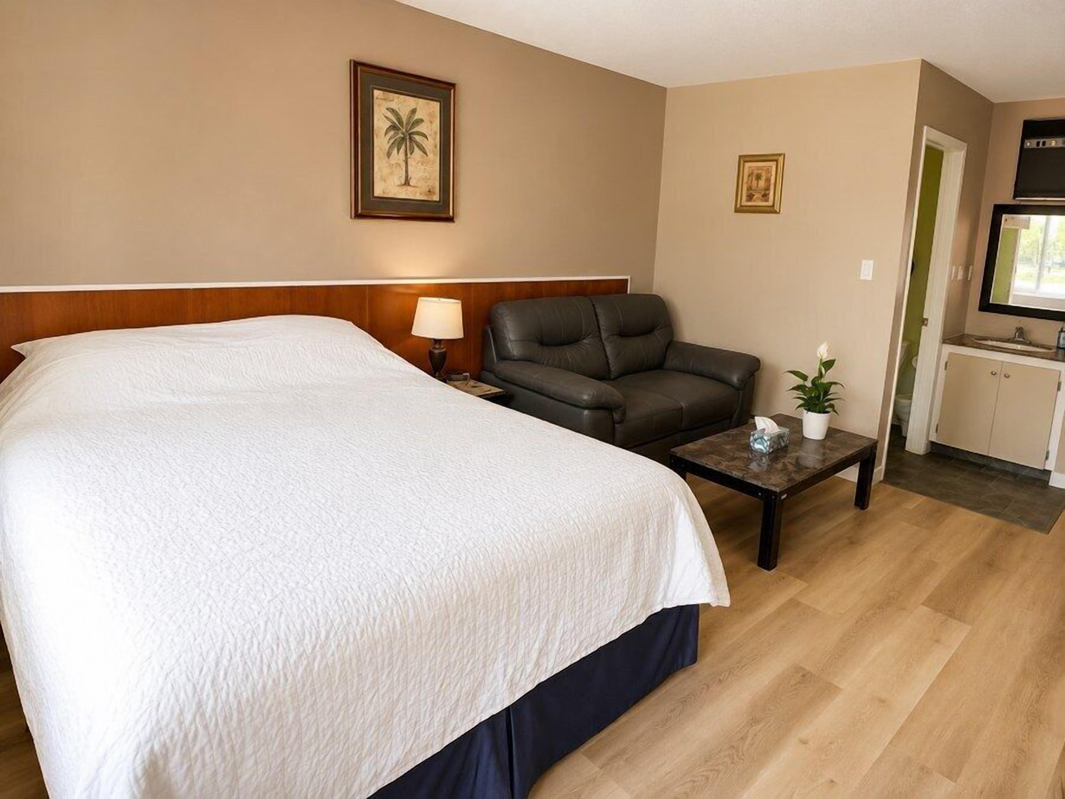 Habitación de hotel con una cama grande, un sillón de cuero, una mesita de noche con lámpara y un área de kitchenette.