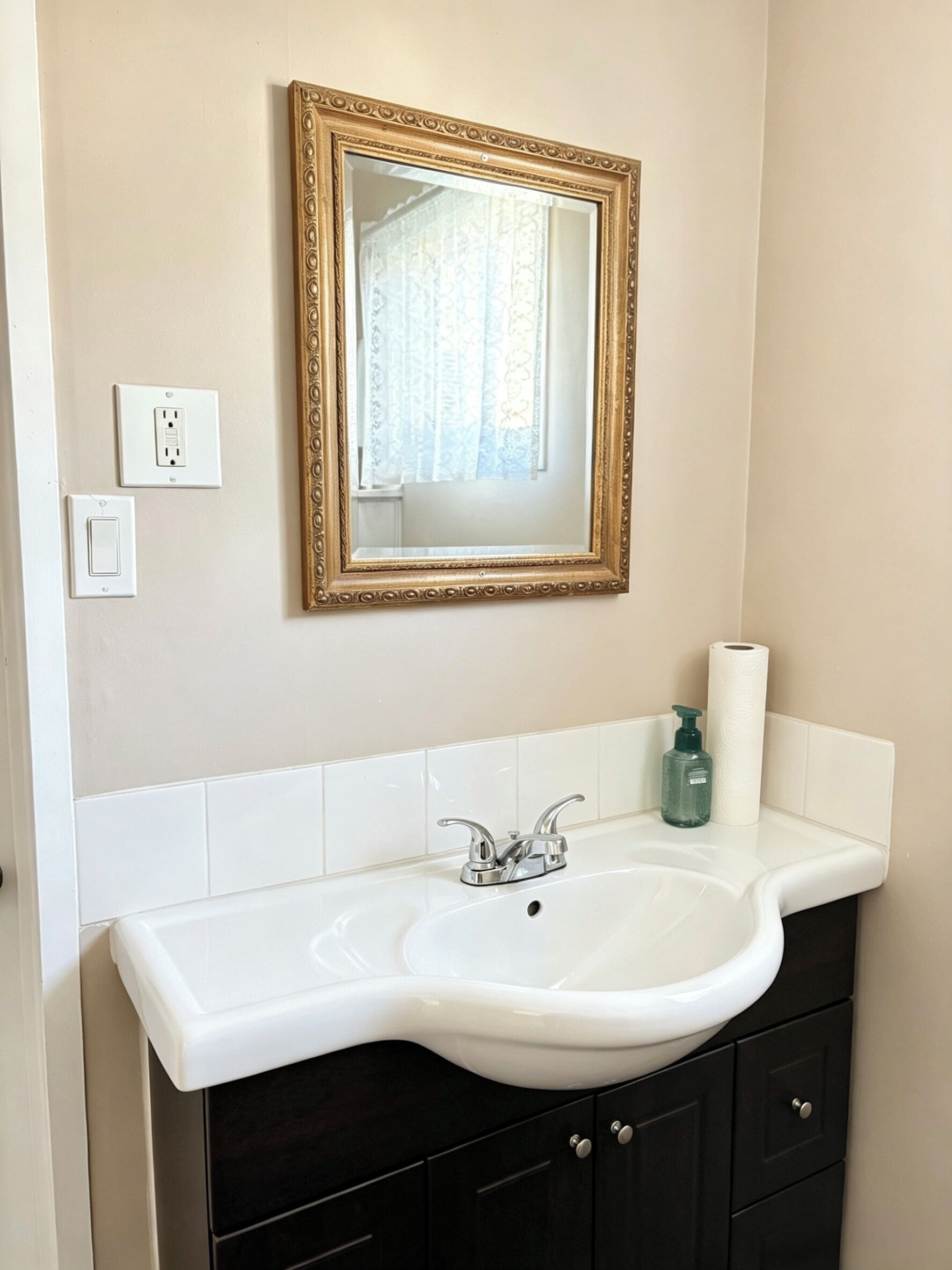 Un baño con un lavamanos blanco, muebles oscuros, un espejo con marco dorado y un dispensador de papel higiénico.