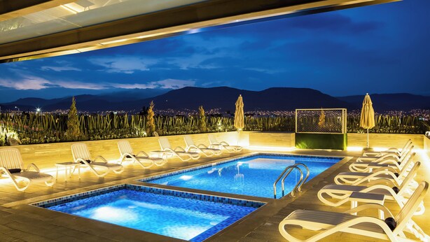 A rooftop pool area with two swimming pools, lounge chairs, and a view of a cityscape at night.