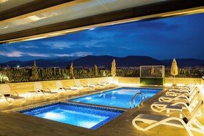 Ein Poolbereich auf dem Dach mit zwei Pools, Liegestühlen und Blick auf eine nächtliche Skyline.