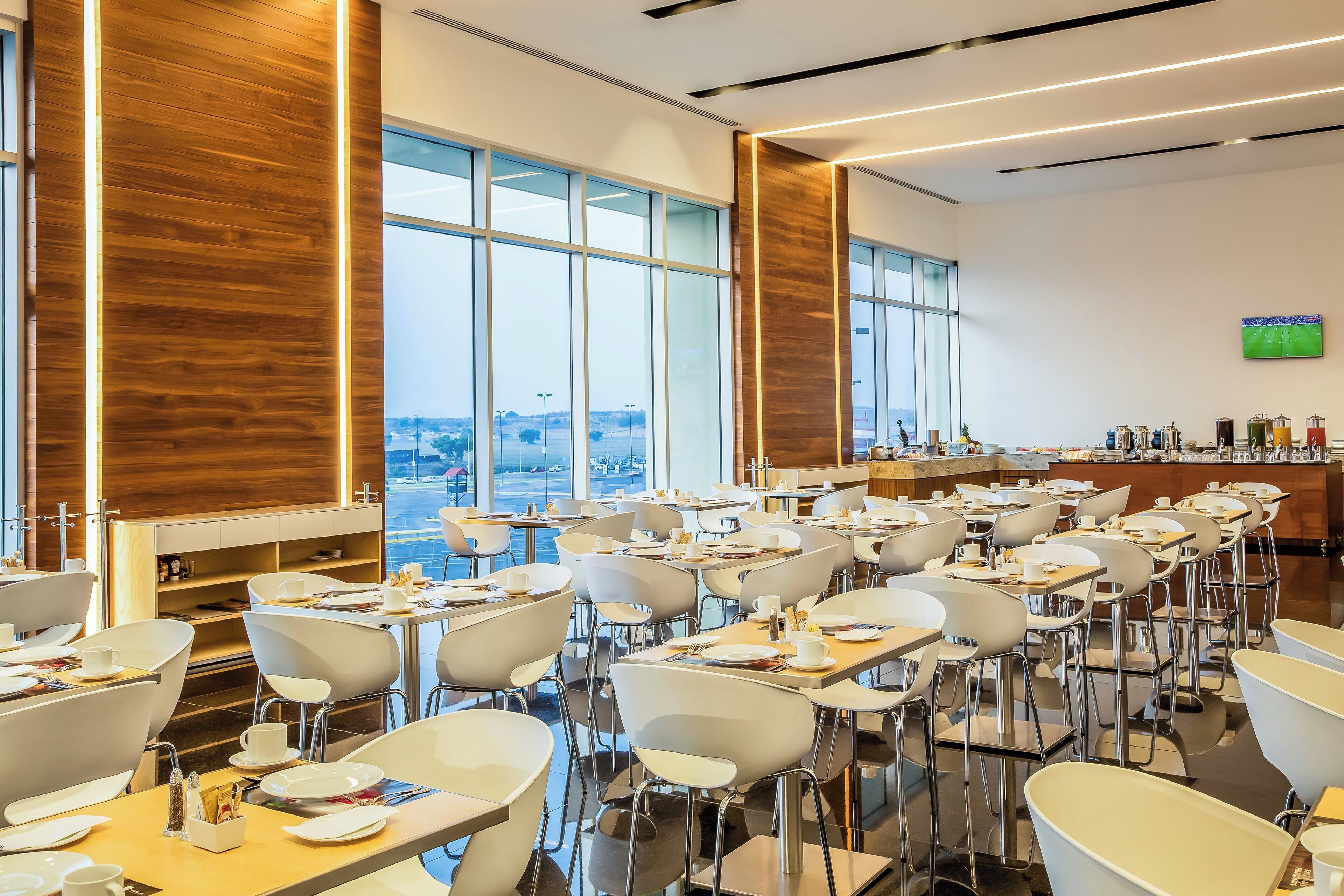 A modern dining area with multiple tables and chairs, large windows offering a view of the city, and a bar area with various amenities.