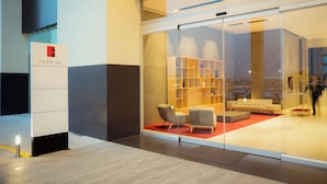 A modern hotel lobby with a glass wall, a seating area with sofas, and a visible bookshelf.