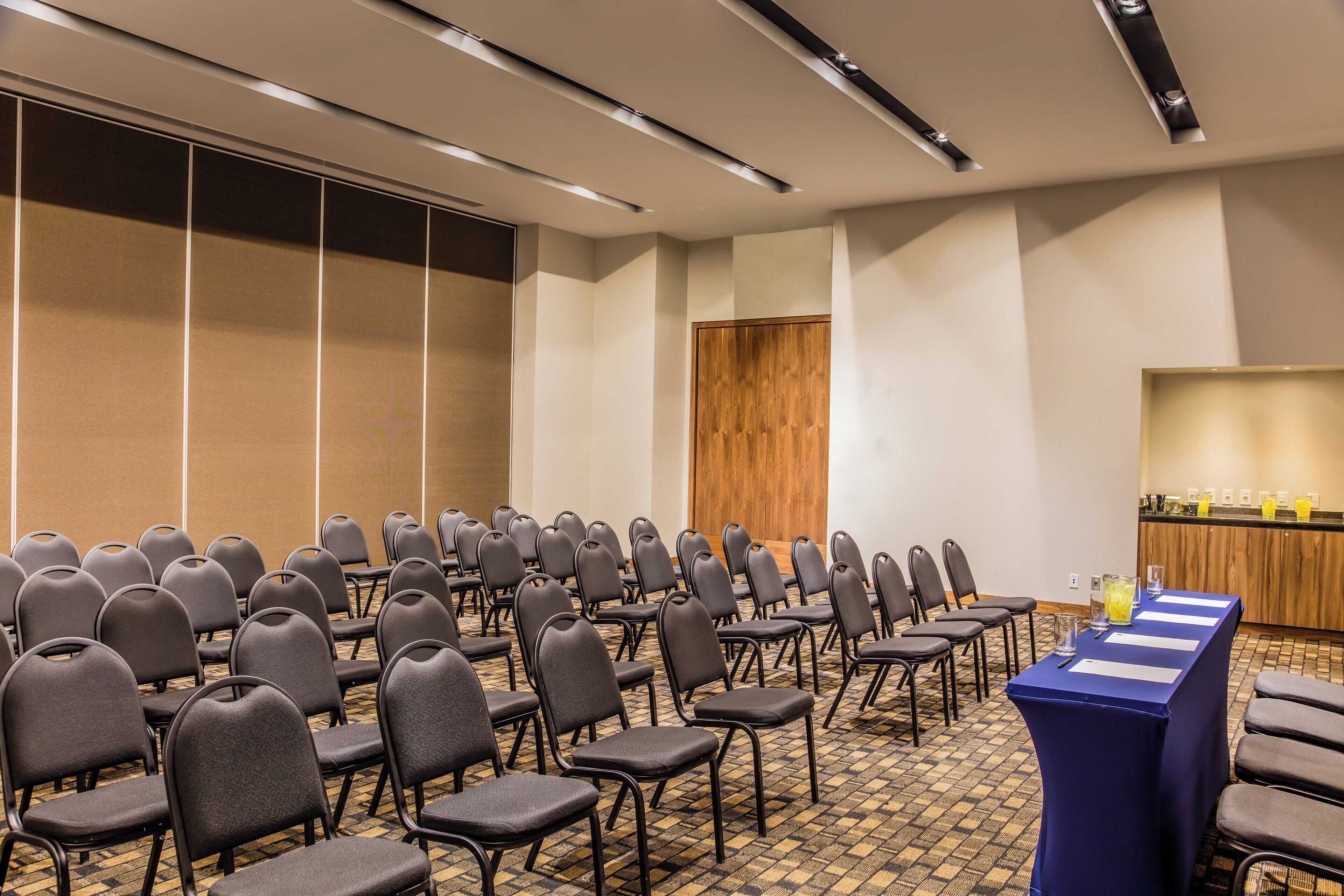 A conference room with rows of chairs, a long table with a blue tablecloth, and a wooden panel wall.