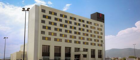 A multi-story hotel with a modern facade, large windows, and a prominent logo on the top right.