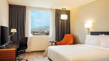Habitación de hotel con cama, escritorio con computadora, silla, lámpara y ventana con cortinas.