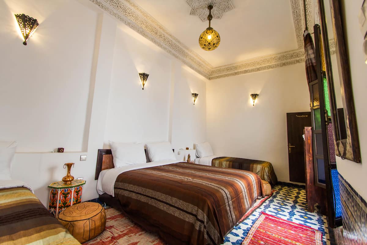 Una habitación de hotel con una cama grande, una mesita auxiliar pequeña, un jarrón decorativo y una alfombra estampada.