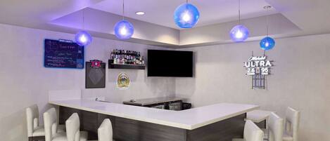 A bar area with a large counter, bar stools, a television, and a neon sign.