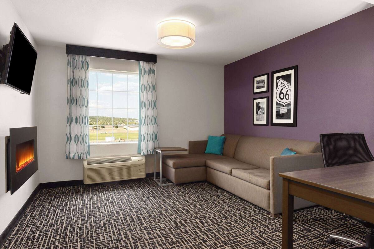 A modern hotel room with a sectional sofa, a small table, a flat-screen TV, a fireplace, and framed pictures on the wall.