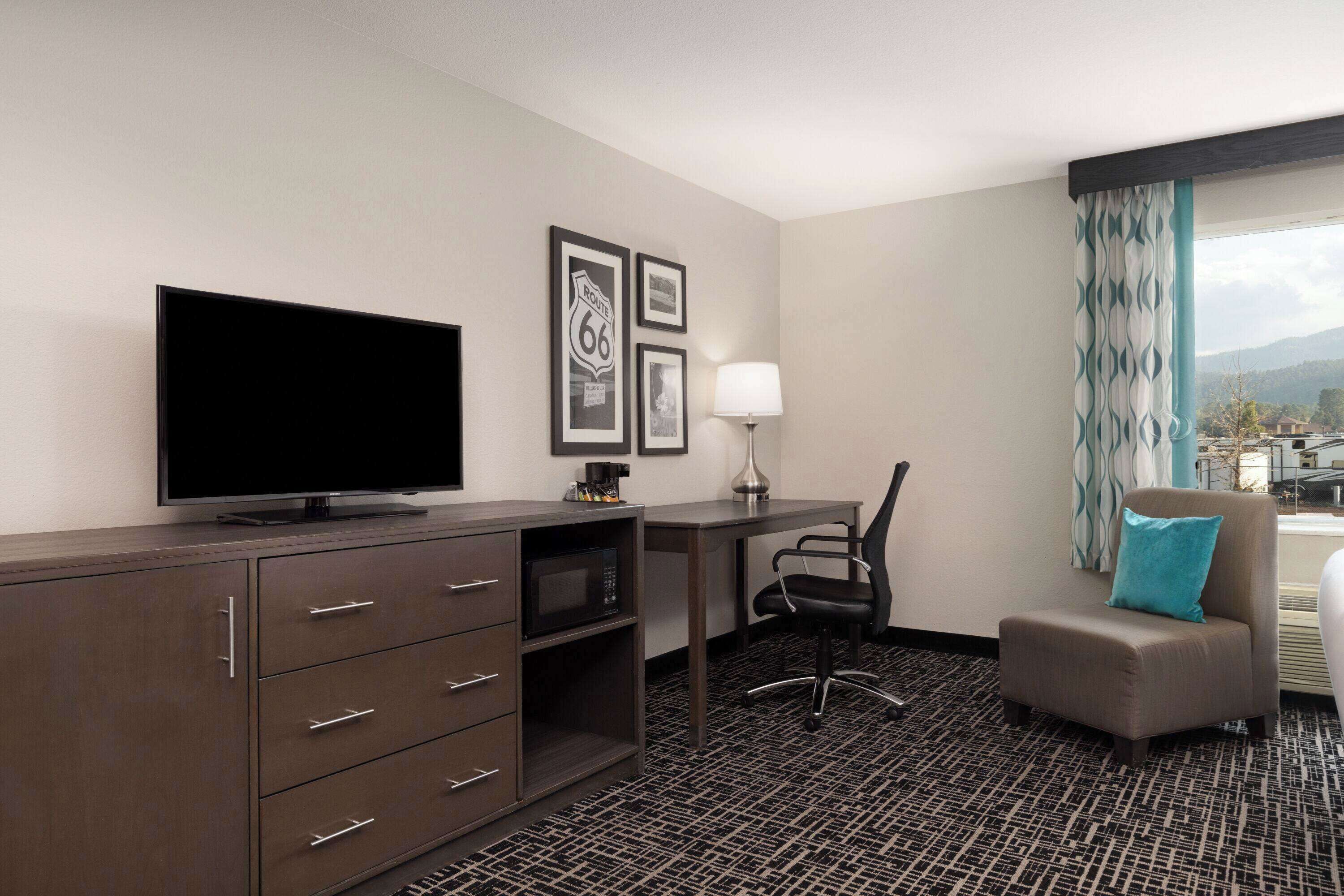 Ein Hotelzimmer mit Flachbildfernseher, Schreibtisch mit Lampe, Stuhl, Sofa mit Kissen und gerahmten Bildern an der Wand.