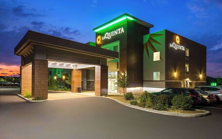 A hotel with a modern design, featuring a prominent 'La Quinta' sign, a covered entrance, and a parking area with several cars.