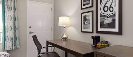 A hotel room with a bed, a desk, a chair, a lamp, a coffee maker, and framed pictures on the wall.