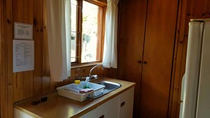 Une petite cuisine équipée avec un plan de travail en bois, un évier et une fenêtre avec des rideaux. Un réfrigérateur blanc se trouve à droite, et un panneau affichant les numéros de téléphone d’urgence est accroché au mur.