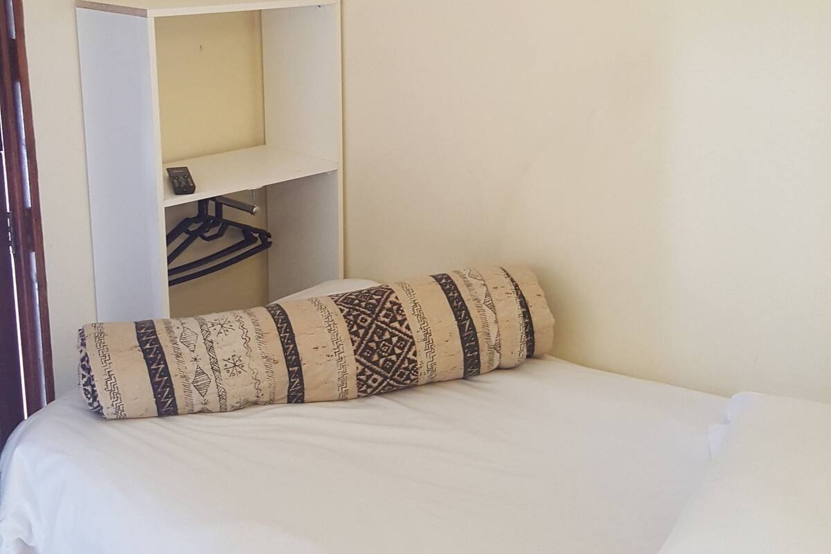 Une chambre à coucher avec un lit, un climatiseur fixé au mur, une étagère avec un livre, un tableau encadré et un porte-manteau suspendu.