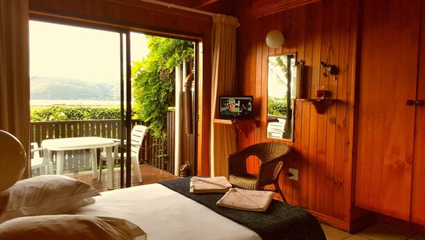 Une chambre dotée d’un lit, d’une télévision, d’un fauteuil en osier et d’un balcon offrant une vue sur la verdure et les montagnes.