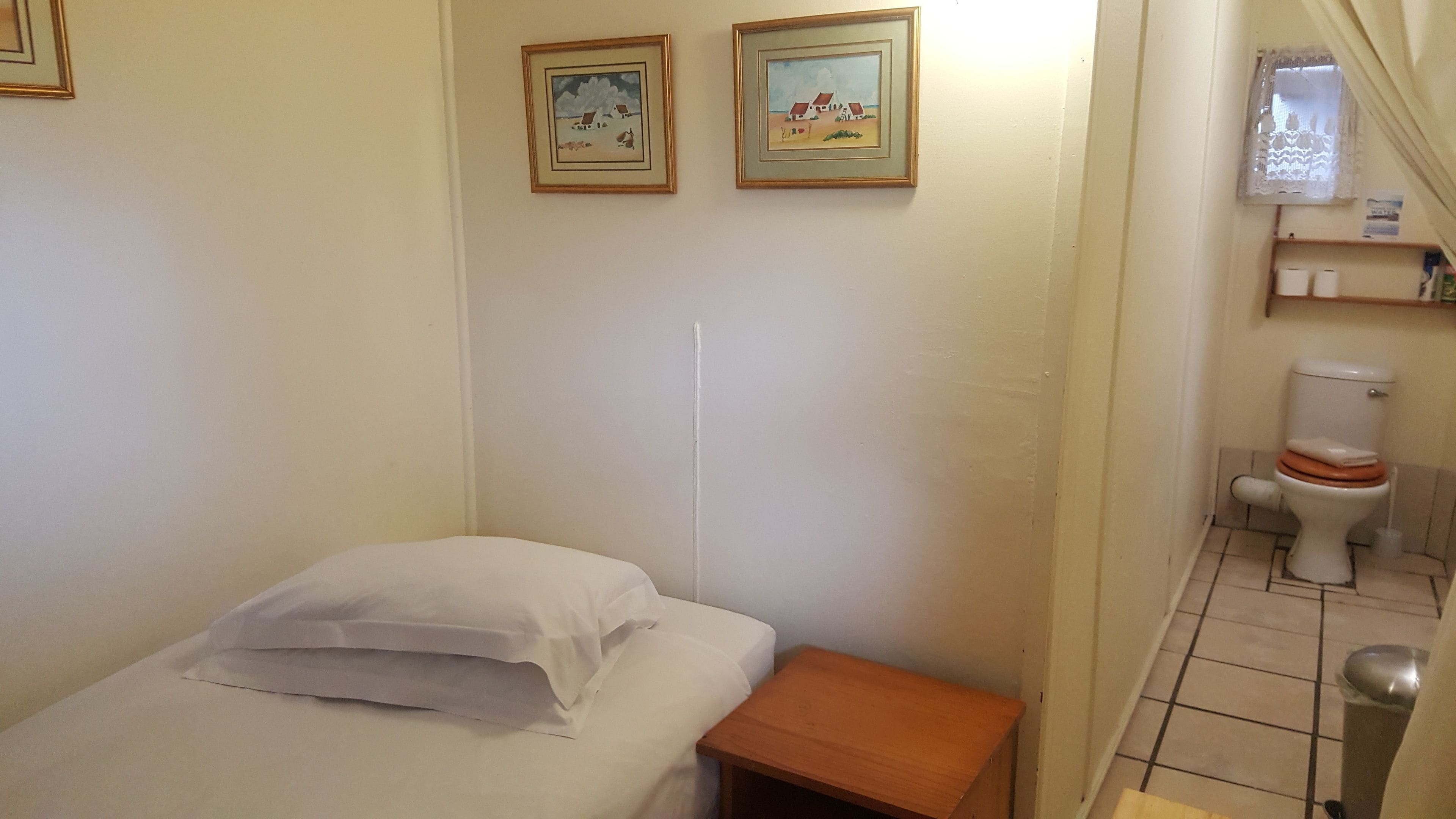 A small, clean room with a bed, a wooden nightstand, a toilet, and two framed pictures on the wall.