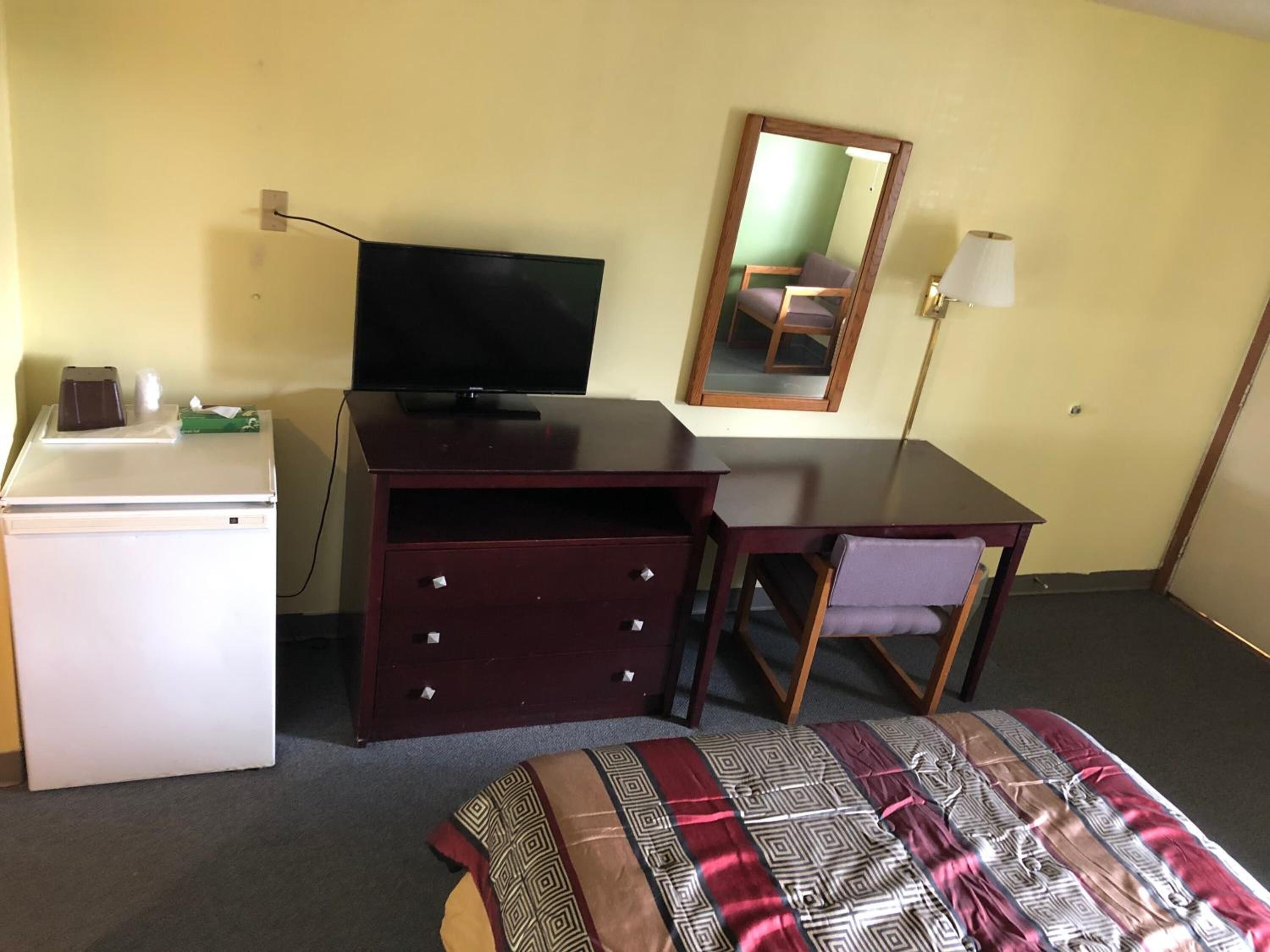 Een kleine kamer met een bed, bureau, televisie, kleine koelkast en spiegel.
