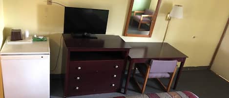 Una habitación compacta con cama, escritorio, televisor, refrigerador pequeño y espejo.