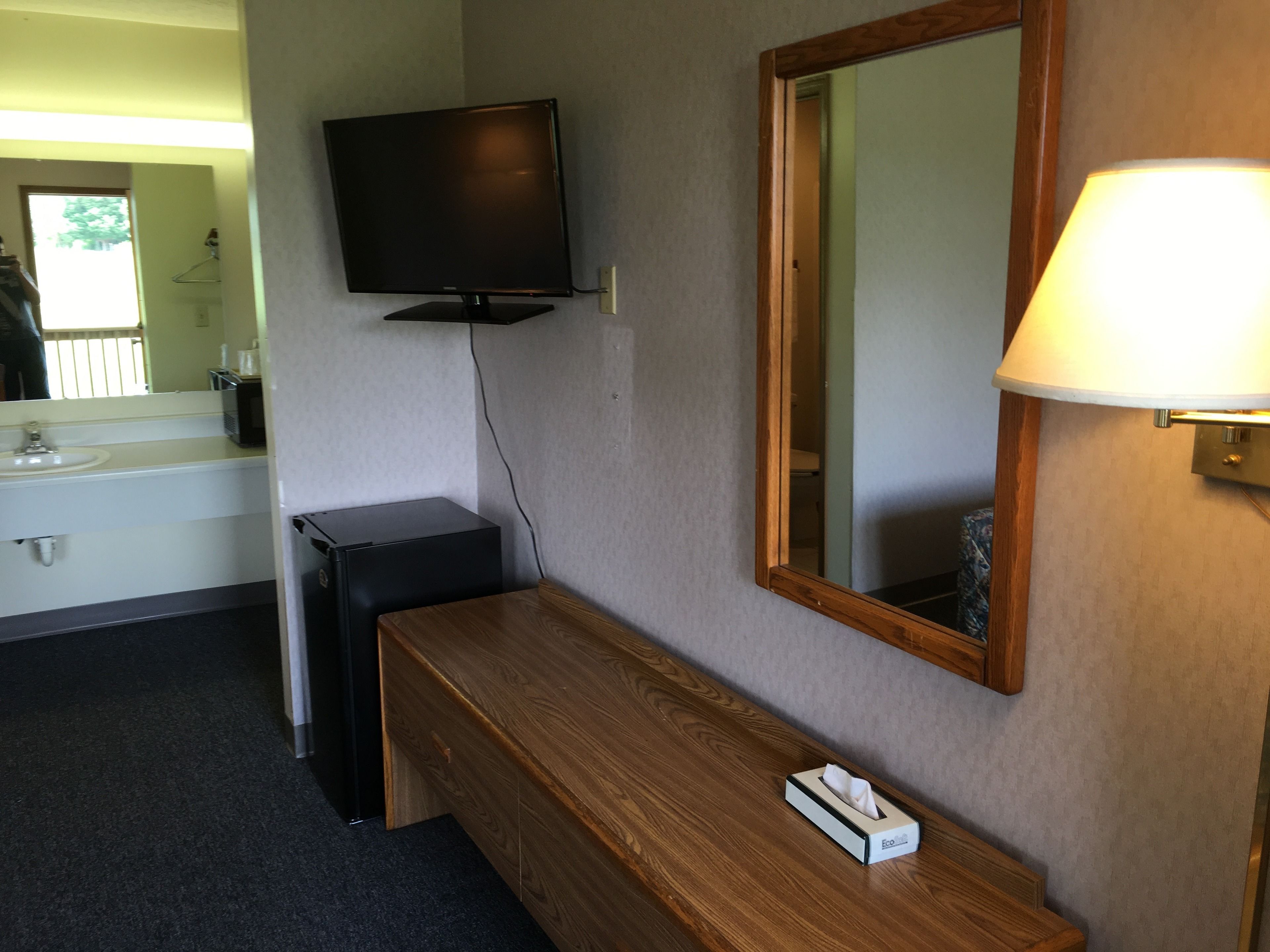 A hotel room with a wooden dresser, a television mounted on the wall, a mirror, and a bedside lamp.