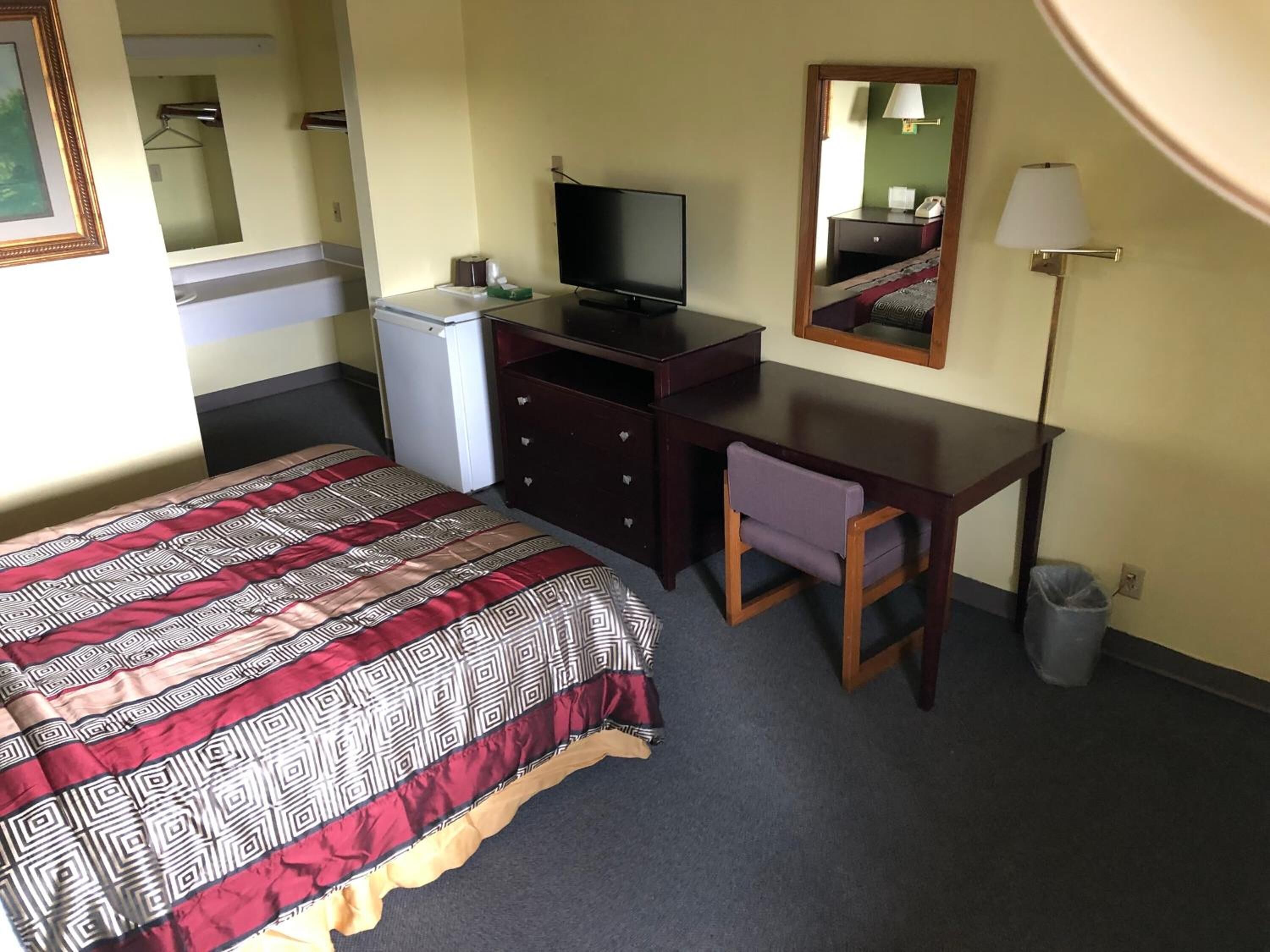 A hotel room with a bed, a desk, a television, a small refrigerator, a chair, a mirror, and a lamp.