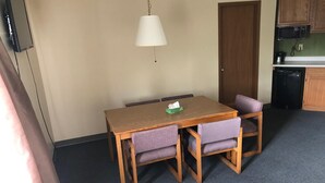 A small dining area with a wooden table and four chairs, a hanging light fixture, and a kitchenette in the background.