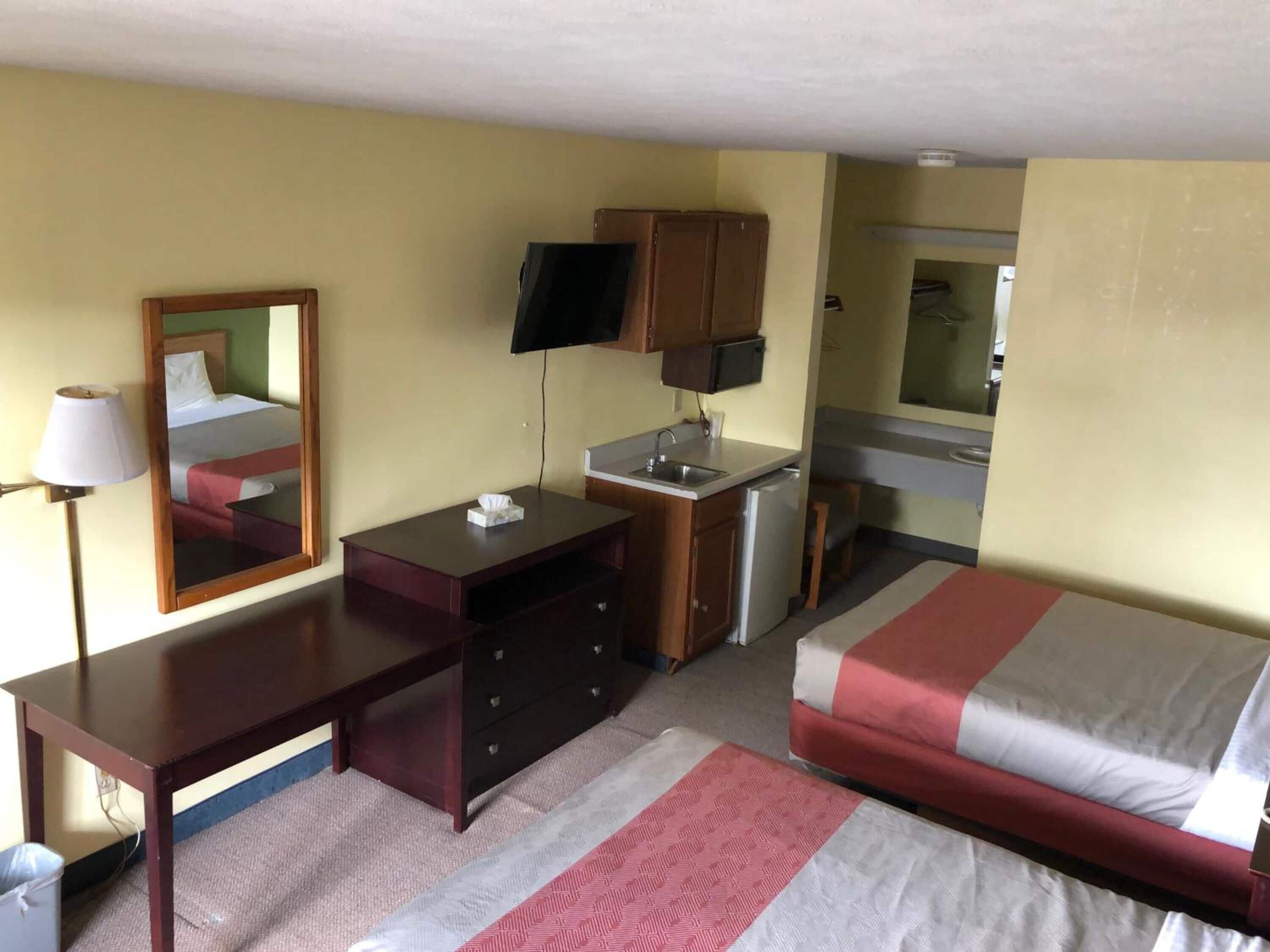 Habitación de hotel compacta con cama, escritorio, espejo, televisor, kitchenette pequeña y baño.