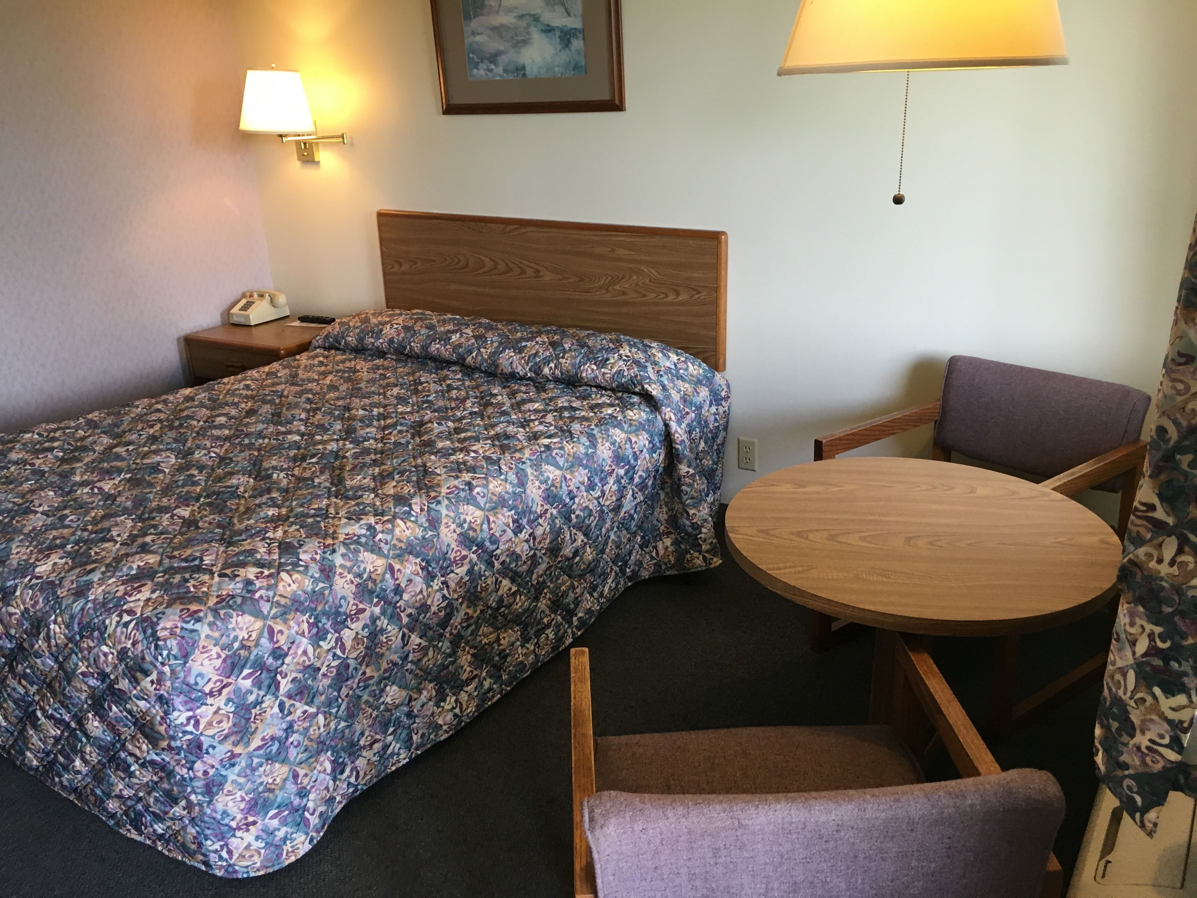 A hotel room with a bed, a round wooden table, a chair, a nightstand with a lamp, and a wall-mounted picture.