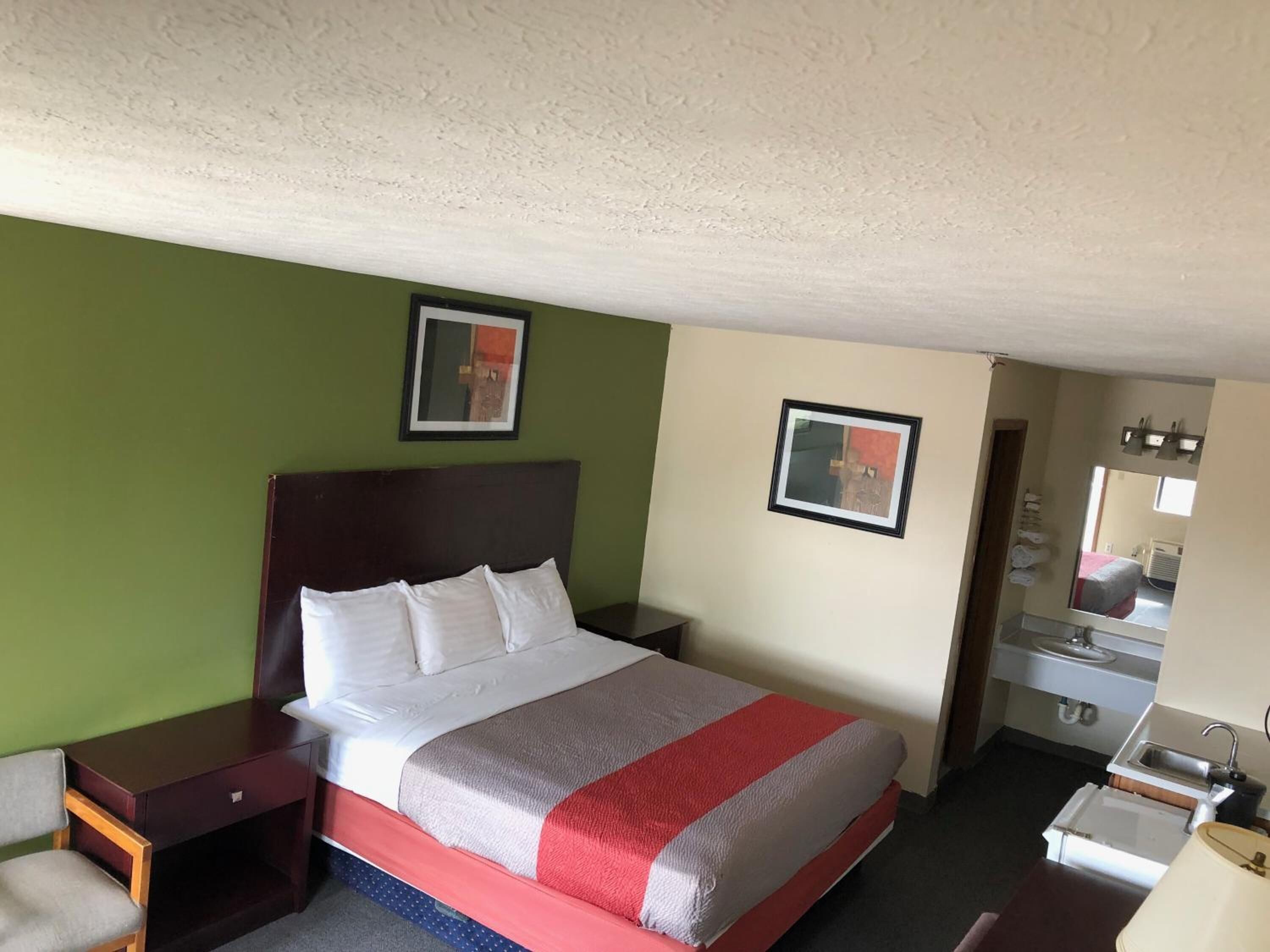 A hotel room with a bed, a nightstand, a chair, a desk, a mirror, and a sink.