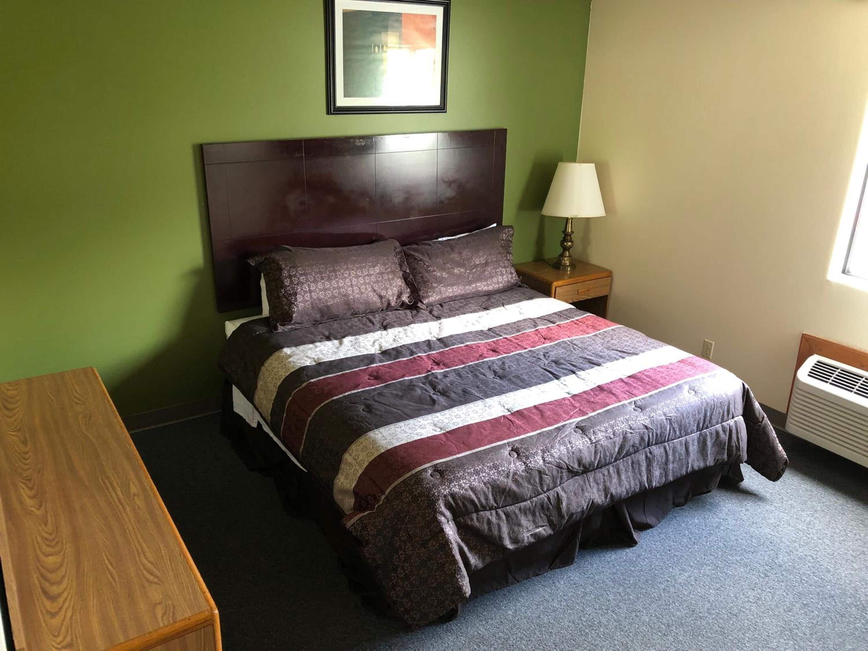 Quarto com cama, mesa de cabeceira, luminária, um banquinho de madeira e um quadro na parede.