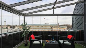 A glass-enclosed balcony with wicker furniture, a small table with a bottle and glasses, and a view of a building with a statue.