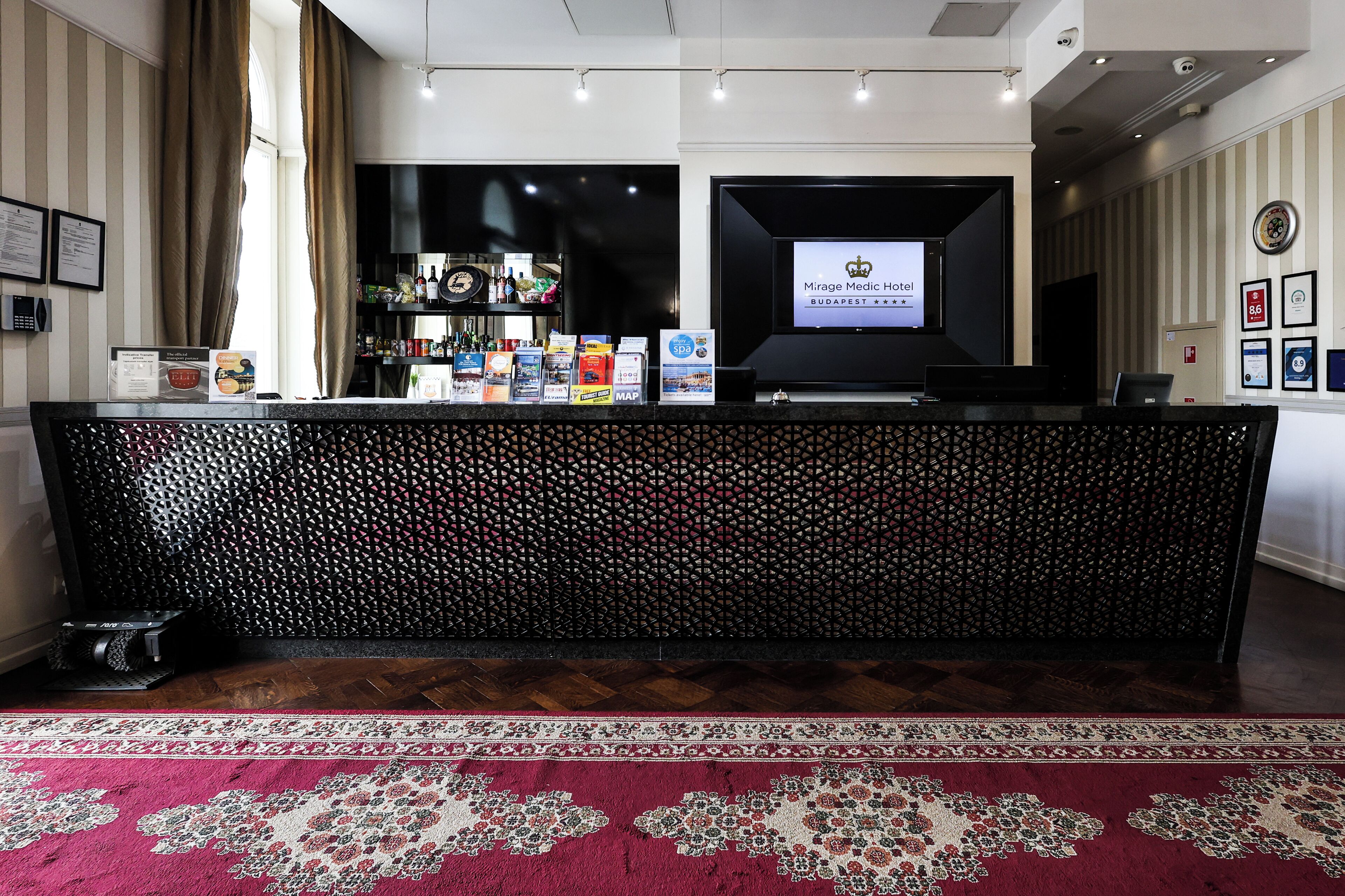 A reception desk with a digital screen displaying the hotel name, a patterned carpet, and a bar area with bottles and snacks.