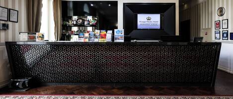 A reception desk with a digital screen displaying the hotel name, a patterned carpet, and a bar area with bottles and snacks.