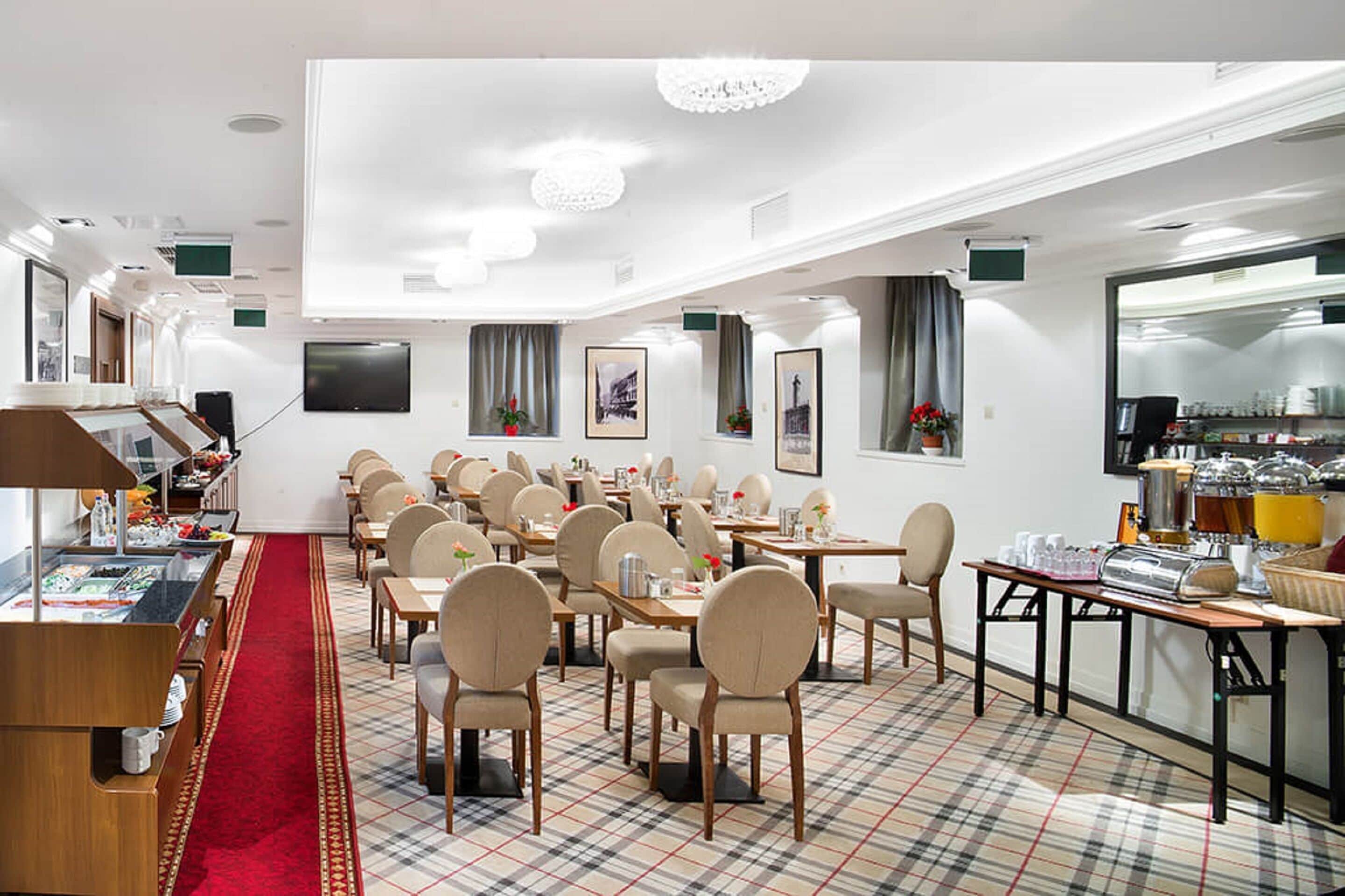 A hotel dining area with multiple tables and chairs, a red carpet, and a kitchen visible through a glass partition.