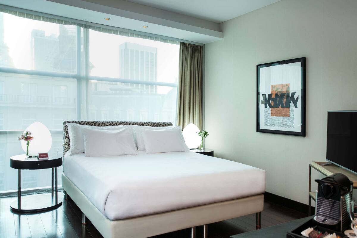 A modern hotel room with a large bed, a flat-screen TV, a small side table with a vase, and a large window offering a city view.