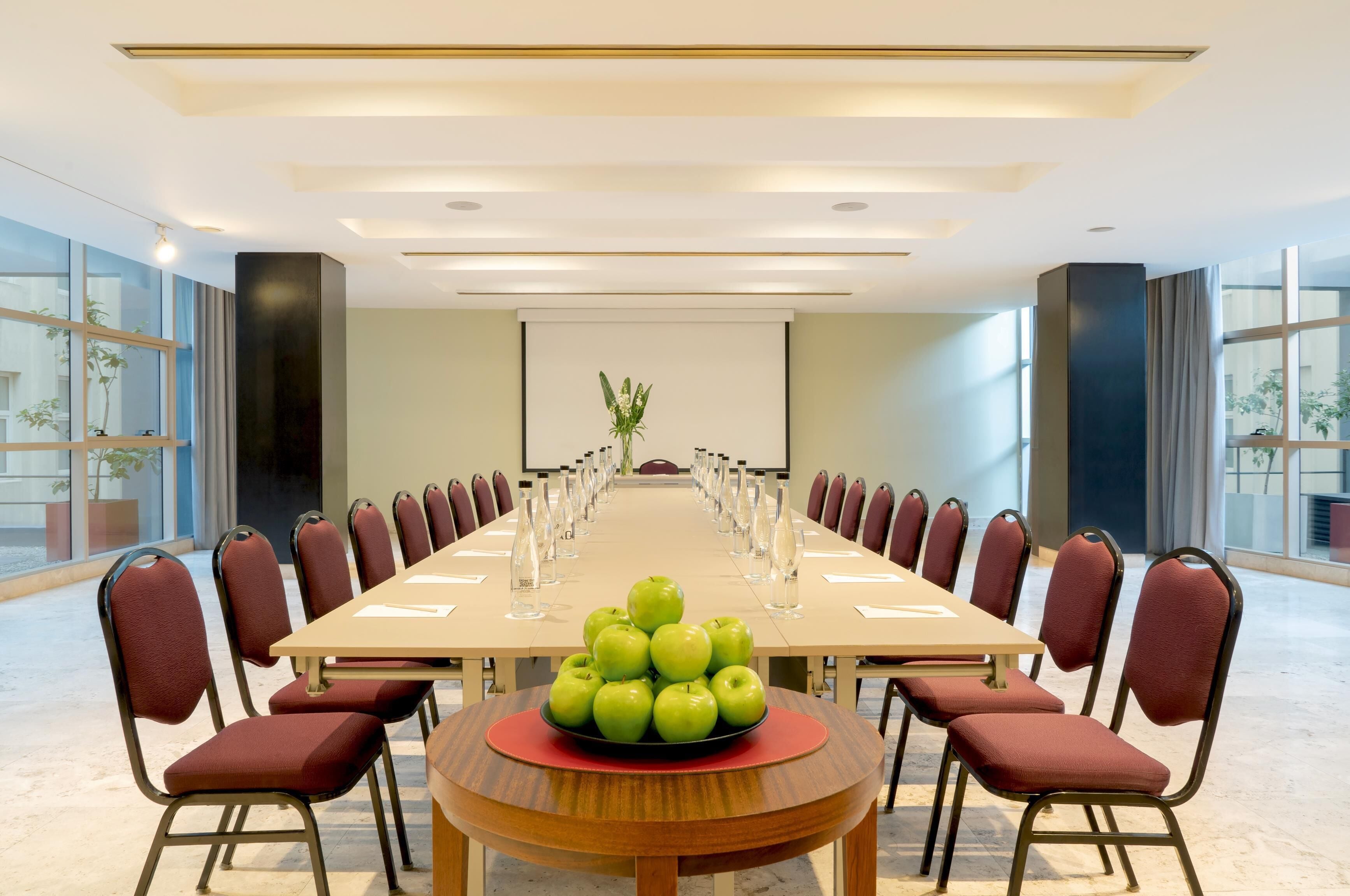 Une salle de conférence avec une longue table, des chaises rouges et un saladier de pommes vertes placé au centre.