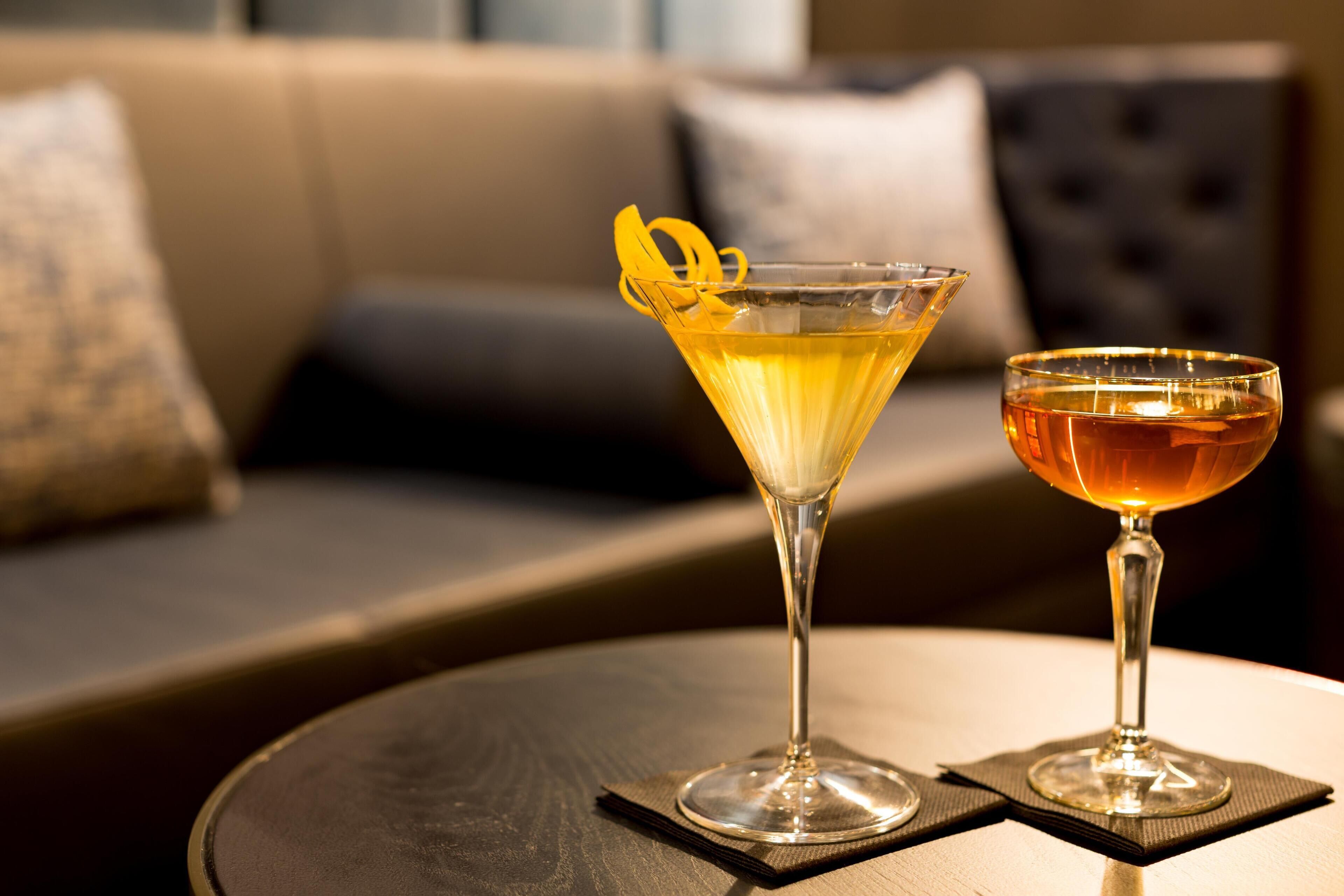 Two cocktails on a round table with a dark coaster, in a room with a sofa and cushions in the background.