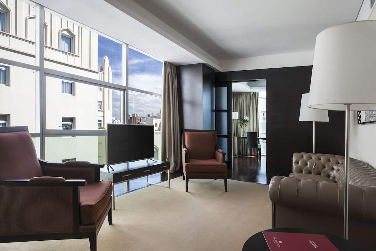 A modern hotel room with a large window, a flat-screen TV, a leather sofa, a brown armchair, and a lamp.