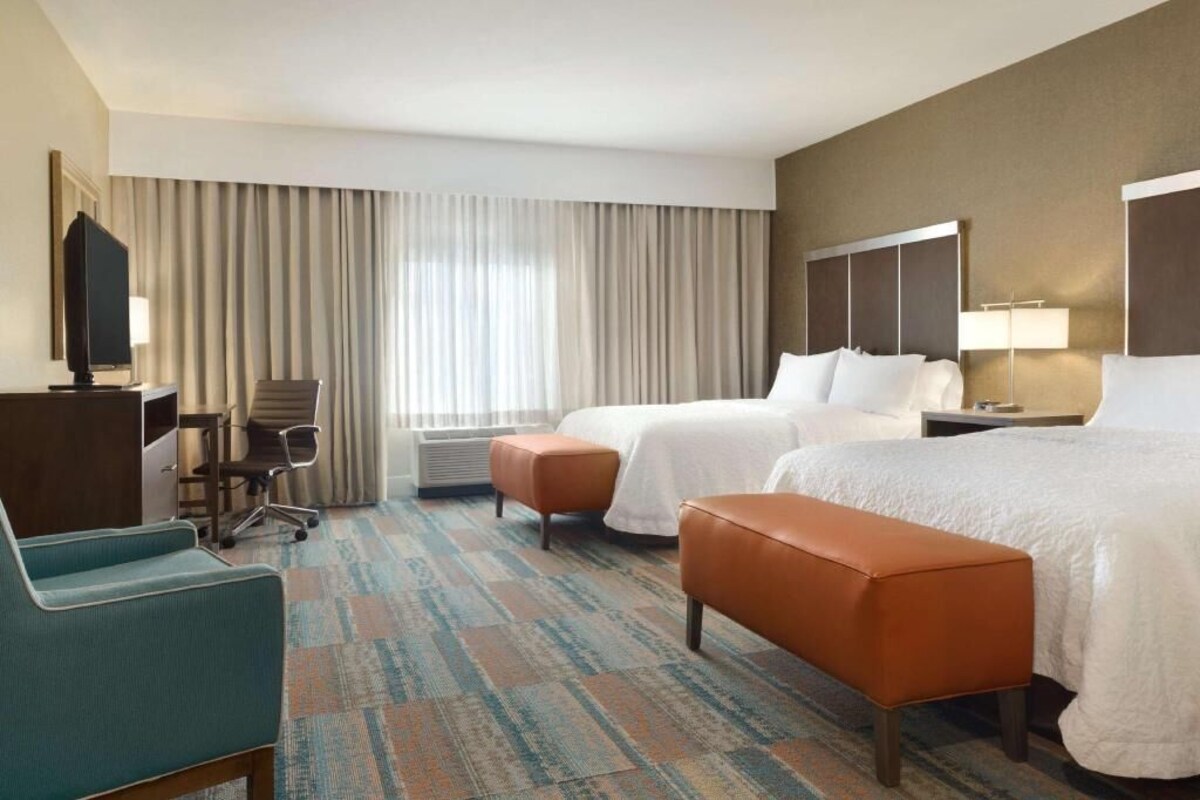 A hotel room with two beds, a desk with a chair, a television, and a large window with curtains.
