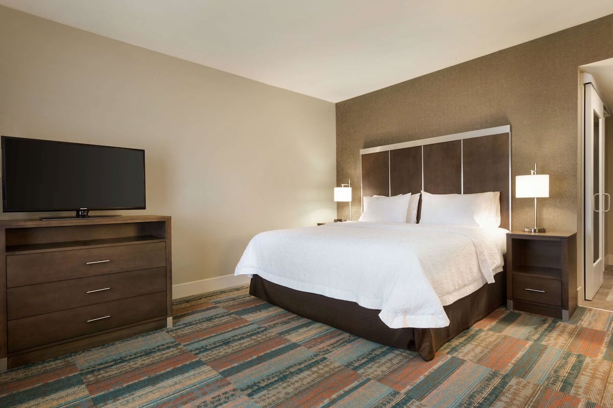 Quarto de hotel com cama, televisão, penteadeira e piso coberto por carpete.