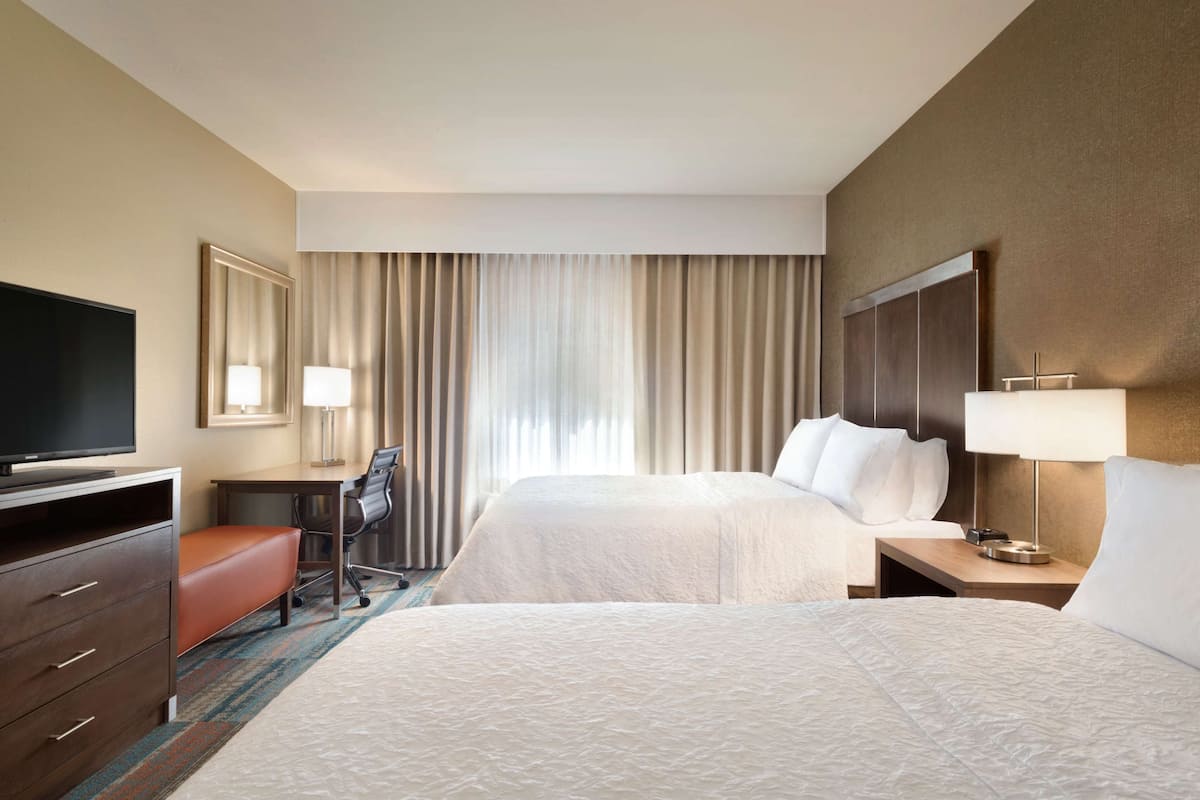 Quarto de hotel com duas camas, uma escrivaninha, uma cadeira, uma televisão e uma janela grande com cortinas.