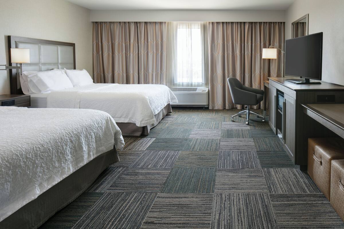 A hotel room with two beds, a desk with a television, a chair, and a large window with curtains.