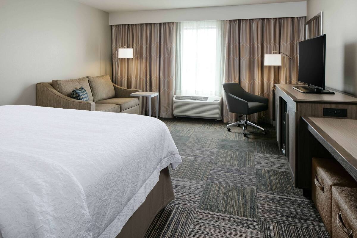 A hotel room with a large bed, a desk with a flat-screen TV, a chair, a small table, and a window with curtains.