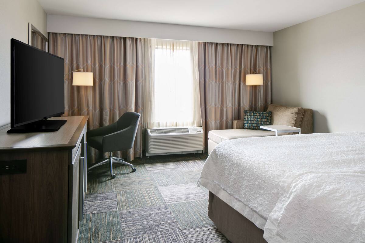 A hotel room with a bed, a desk with a television, a chair, a small table, and a window with curtains.