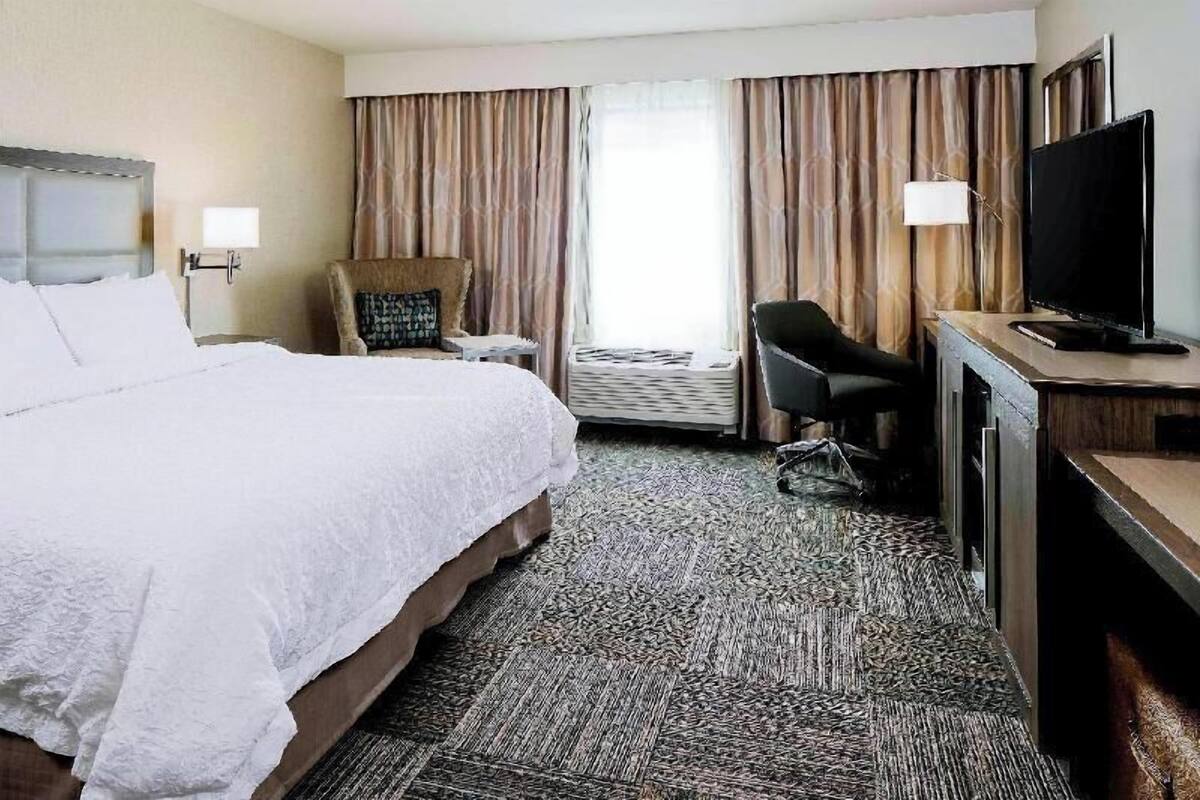 A hotel room with a bed, a desk with a TV, a chair, a lamp, and a window with curtains.