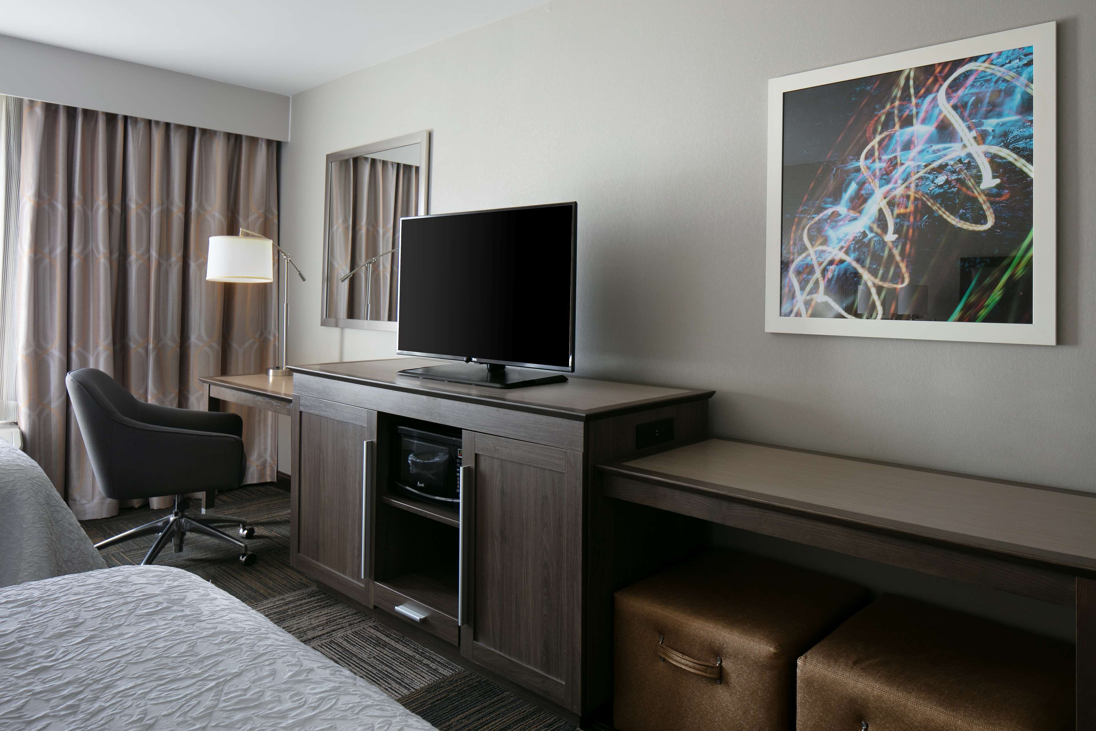 A hotel room with a bed, a desk with a flat-screen TV, a chair, a cabinet, and a large abstract painting on the wall.