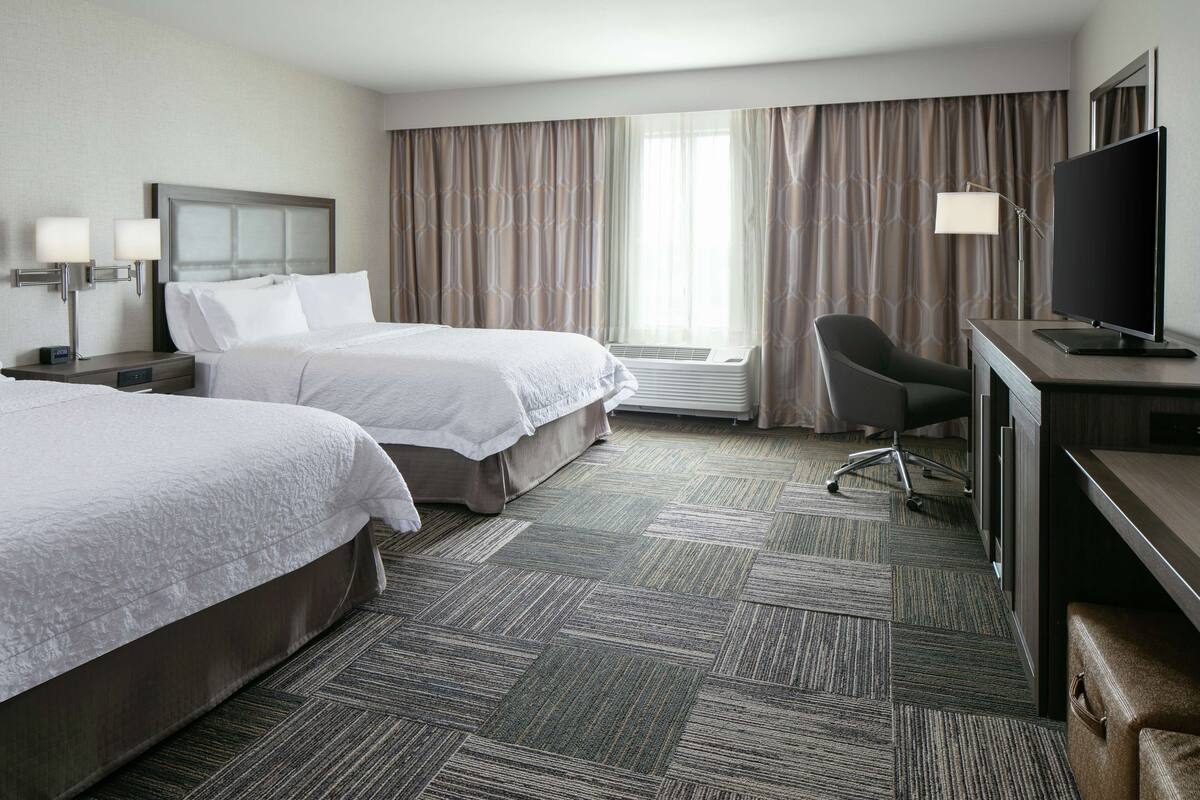 A hotel room with two beds, a desk with a TV, a chair, and a large window with curtains.