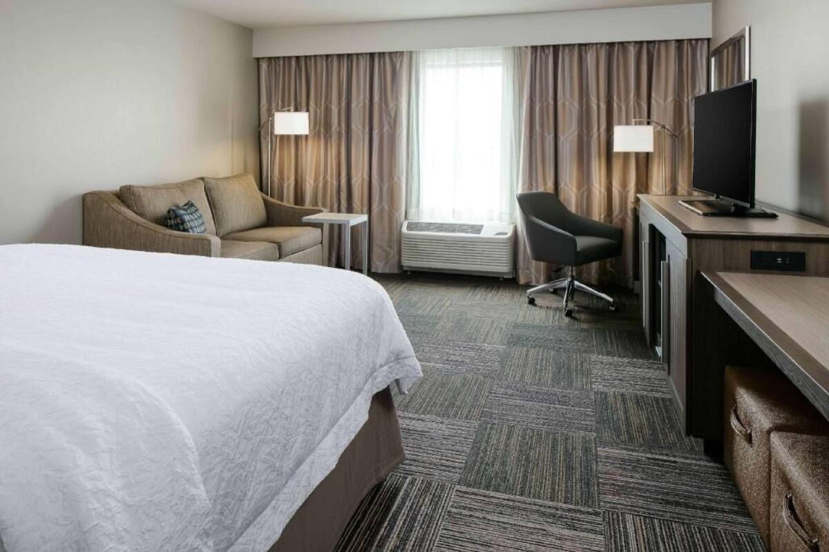 A hotel room with a large bed, a desk with a flat-screen TV, a chair, a small table, and a window with curtains.